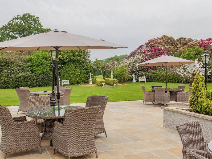 Een terras buiten met rieten meubilair en parasols, omgeven door weelderig groen en bloeiende planten.