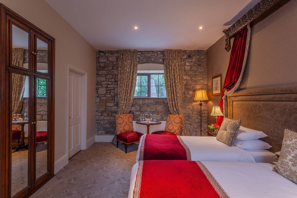 Ein Hotelzimmer mit Steinwand, zwei Betten mit roter und weißer Bettwäsche, einem kleinen Tisch mit Lampe und Blick ins Freie durch ein Fenster.
