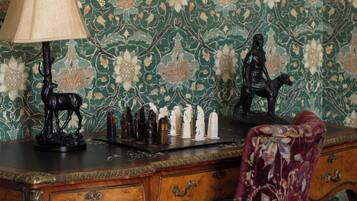 A room with a floral wallpaper, a wooden desk with a lamp, a chair, and a framed mirror.