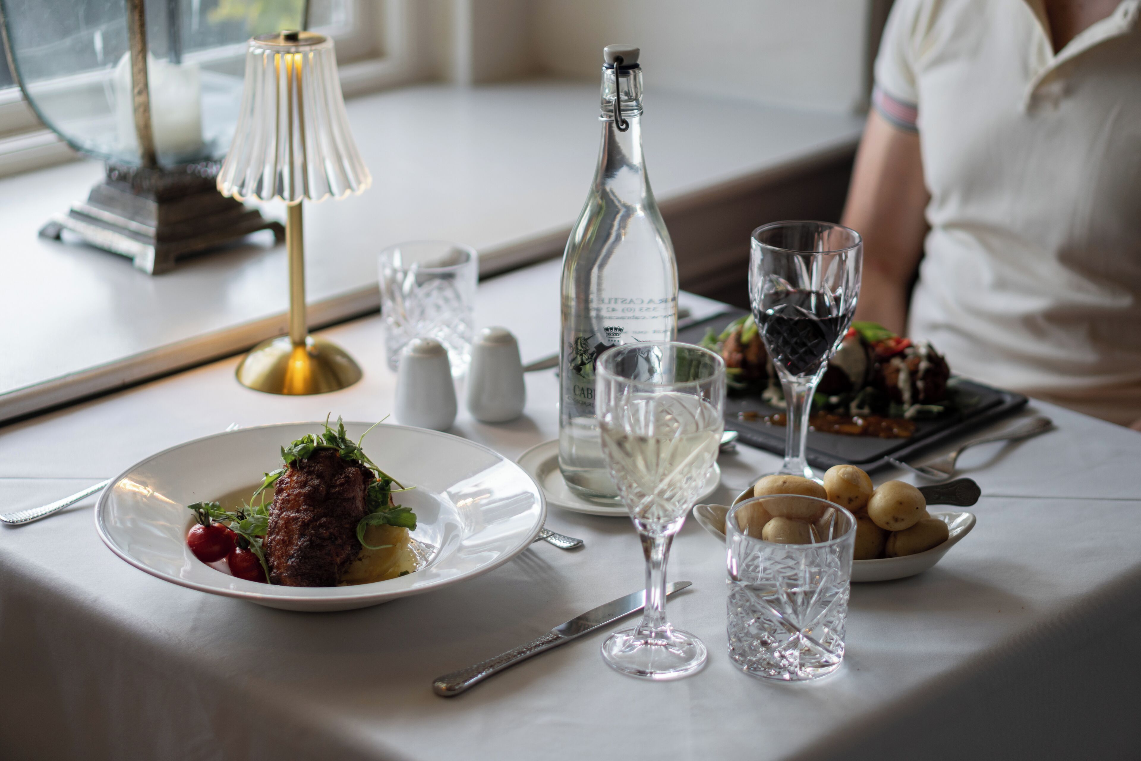 Une table à manger dressée avec une assiette de nourriture, une bouteille, des verres et une lampe.