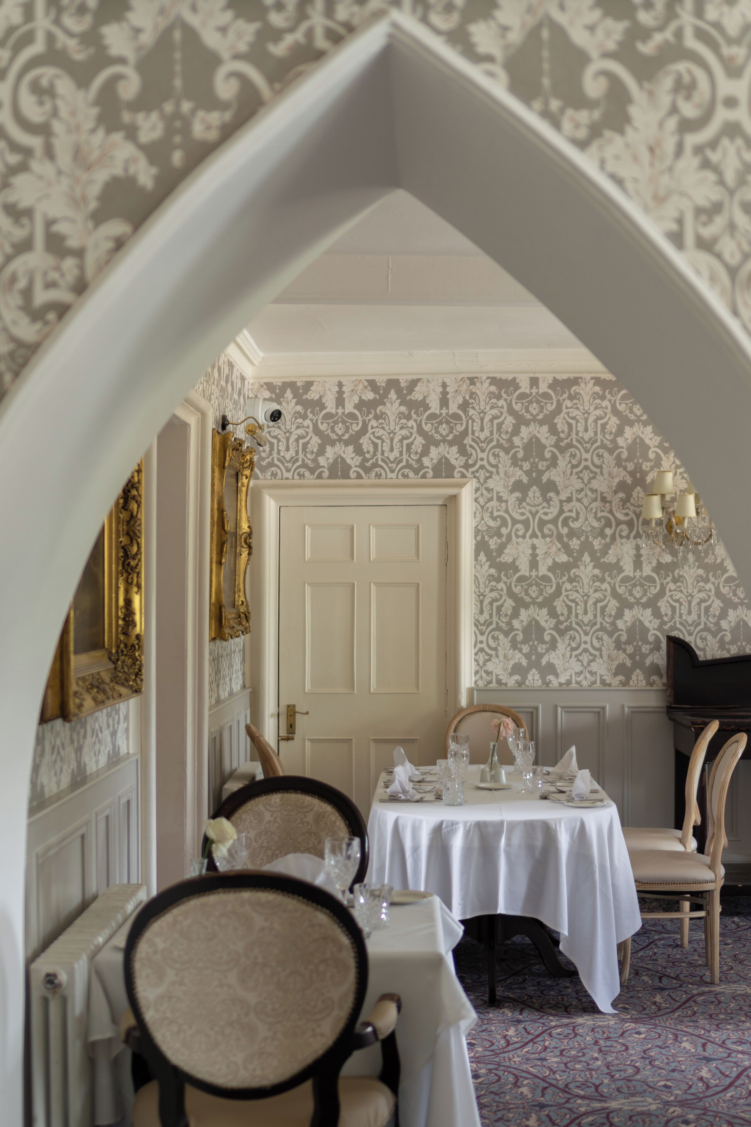 A dining area with a table set for two, a piano, and ornate wallpaper.