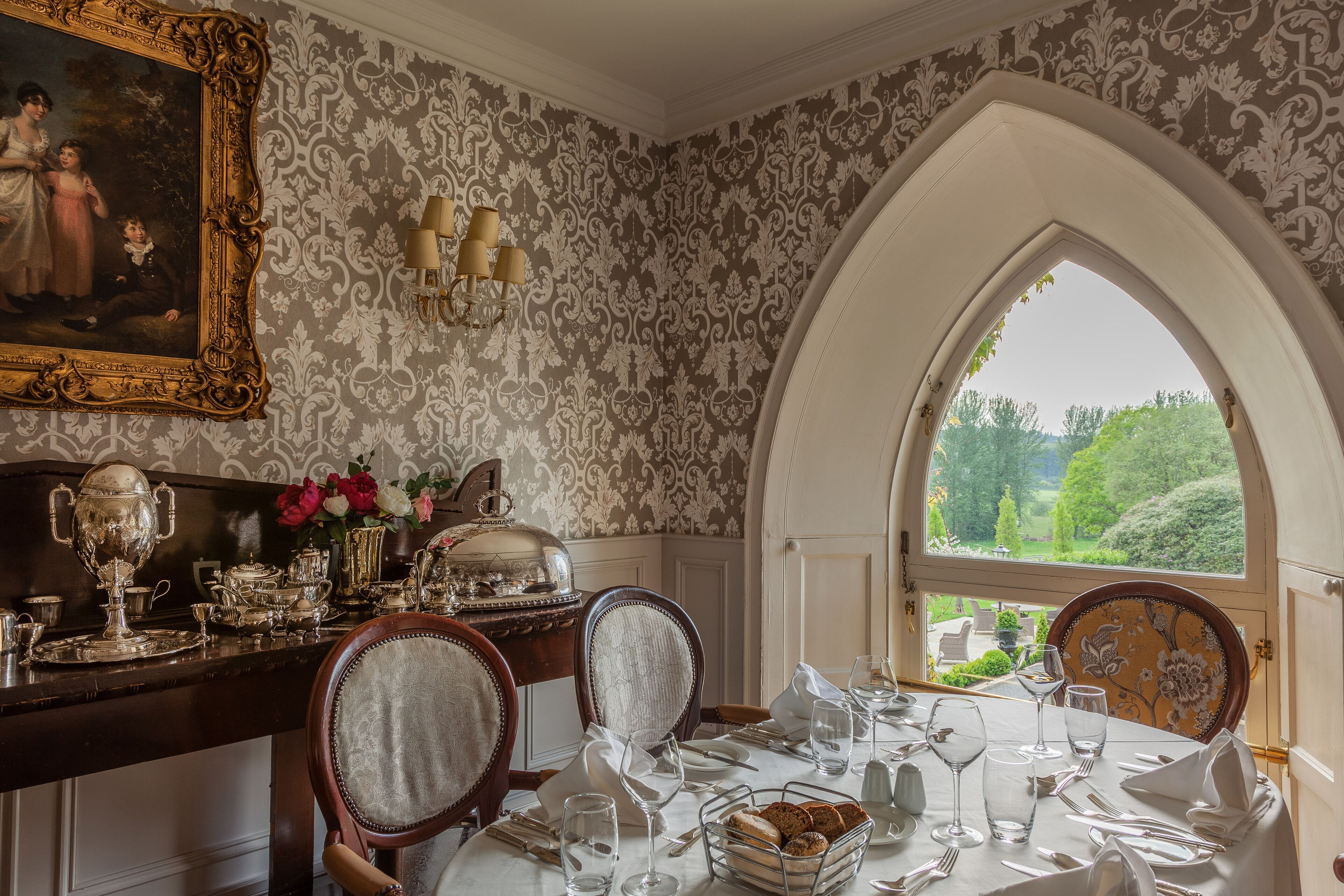 Comedor con una mesa preparada para cuatro personas, un candelabro y vistas a través de un ventanal arqueado.