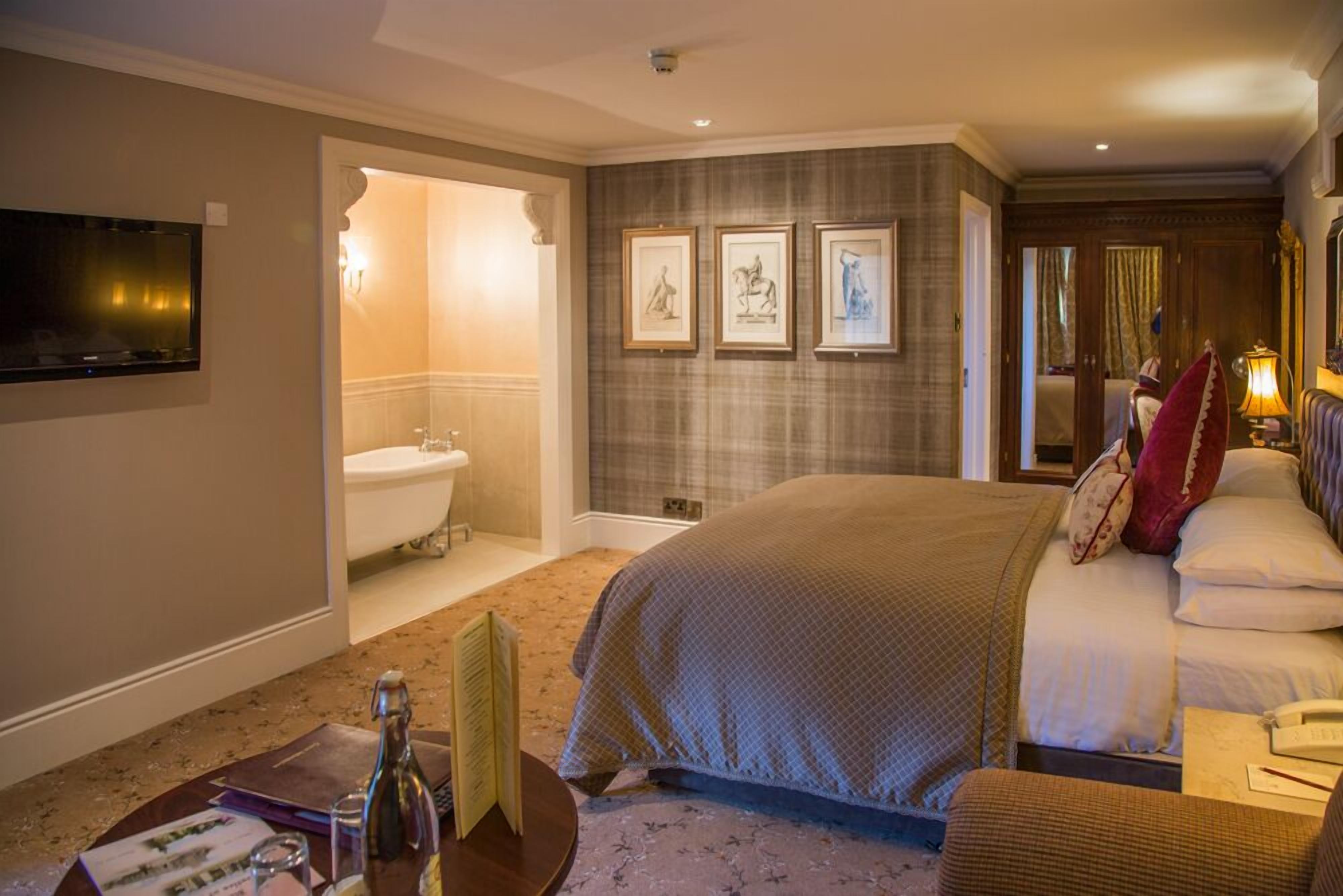 A hotel room with a large bed, a sitting area with a chair, a small table with a bottle and papers, a TV mounted on the wall, and framed artwork on the wall.