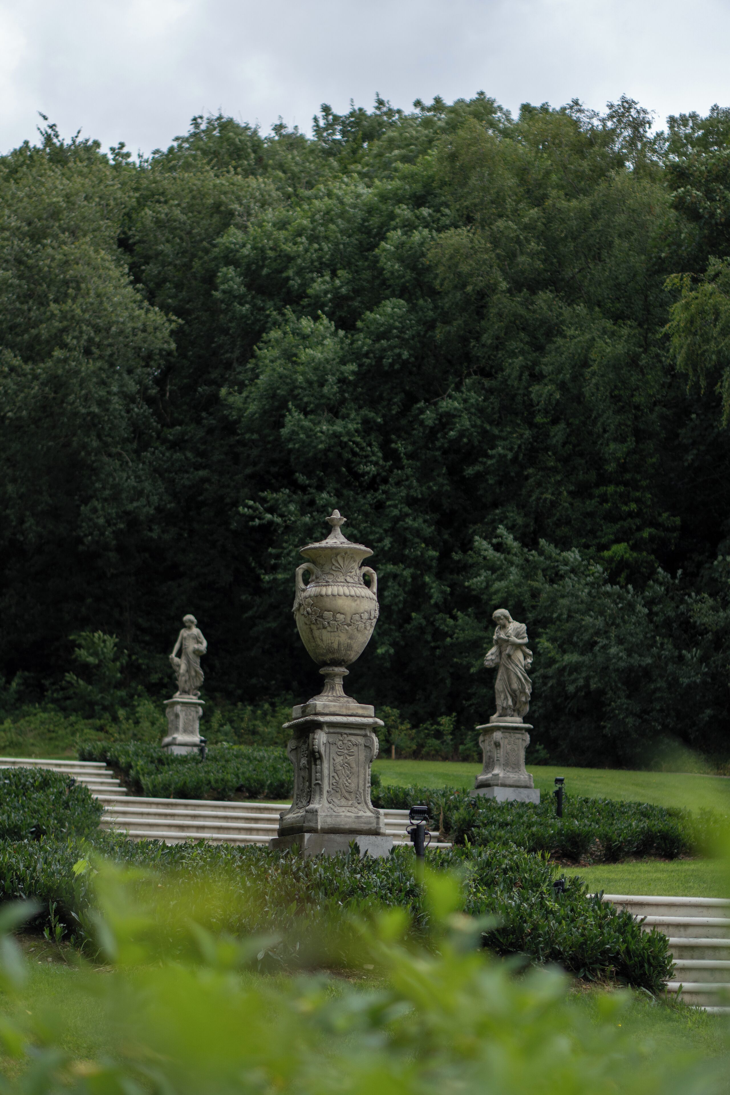 Un jardin agrémenté d’un grand vase et de statues, entouré de haies et d’arbres bien entretenus.