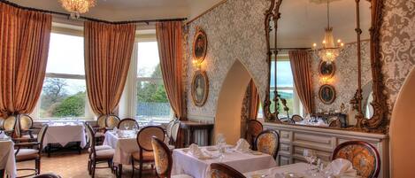 A dining room with tables set for service, ornate wall mirrors, and a chandelier.