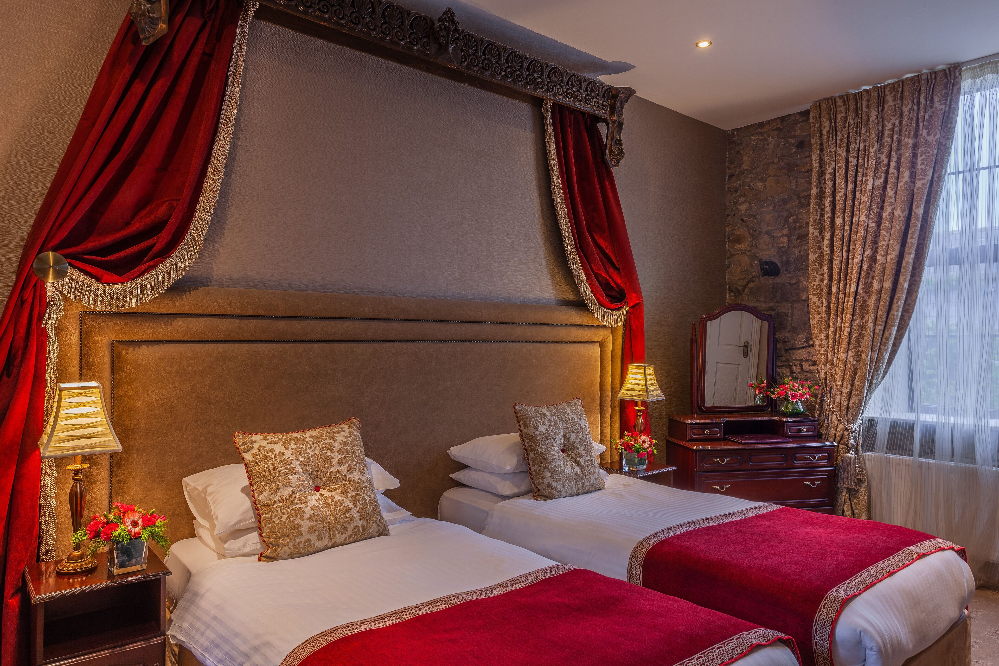 A hotel room with two beds, red curtains, a wooden headboard, a nightstand with a lamp, and a dresser with a mirror.