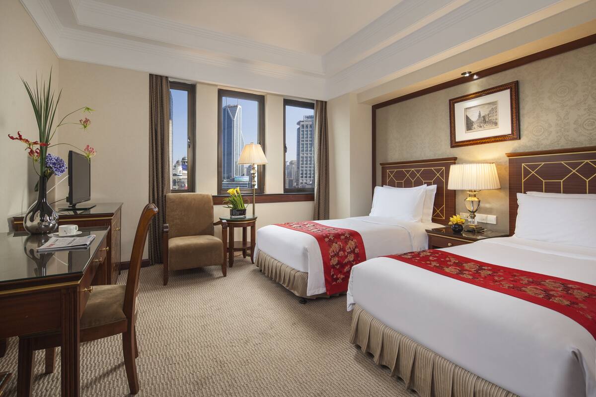 Habitación de hotel con dos camas, un escritorio, una silla, una mesita con una lámpara y una ventana con vistas a la ciudad.