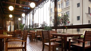 Un terrazzo in legno con tavoli e sedie, ampie finestre con piante e vista sugli edifici.