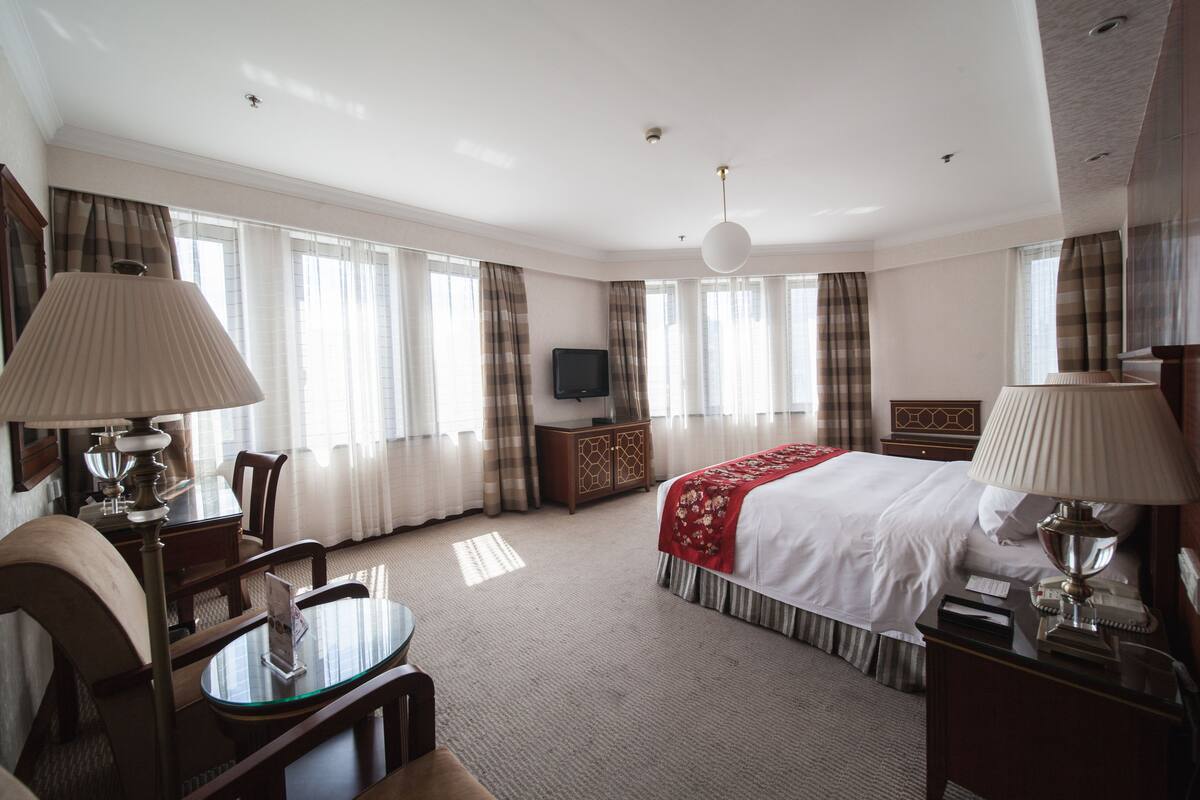 A hotel room with a bed, bedside table, lamp, TV, and a desk with a chair.