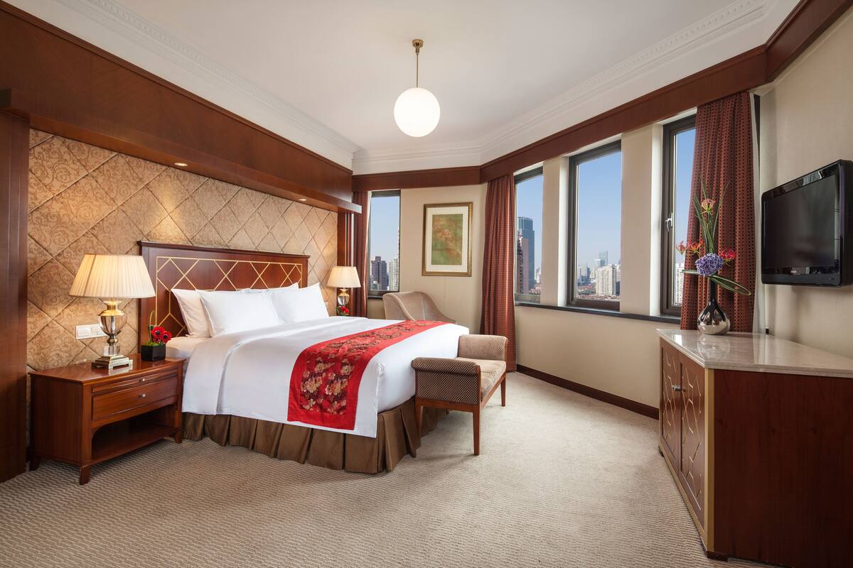 Habitación de hotel con una cama grande, un escritorio con lámpara, una silla, televisor y vistas a la ciudad a través de amplias ventanas.