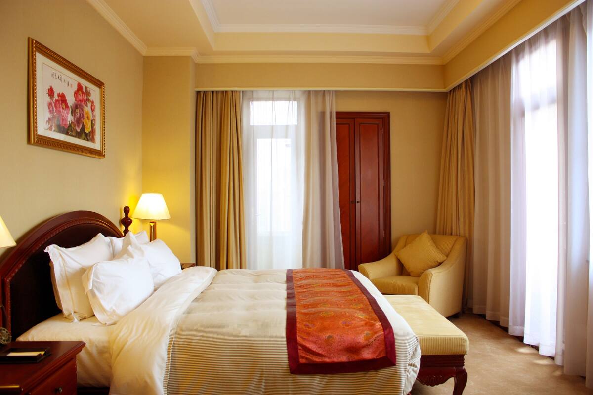 A hotel room with a bed, bedside table, lamp, chair, and curtains.