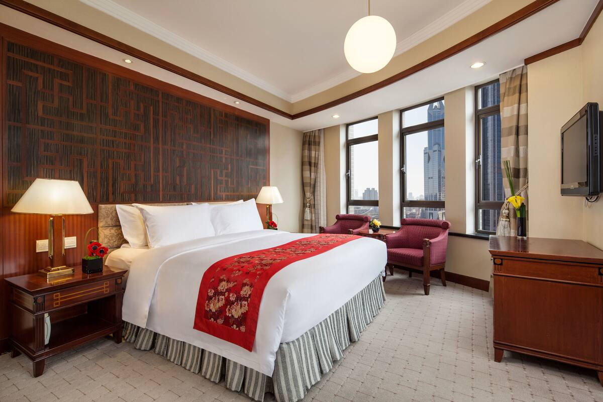 Habitación de hotel con una cama grande, dos lámparas de noche, una colcha decorativa roja, una mesita de noche de madera, un aparador de madera, un sillón rojo y un ventanal con vistas a la ciudad.