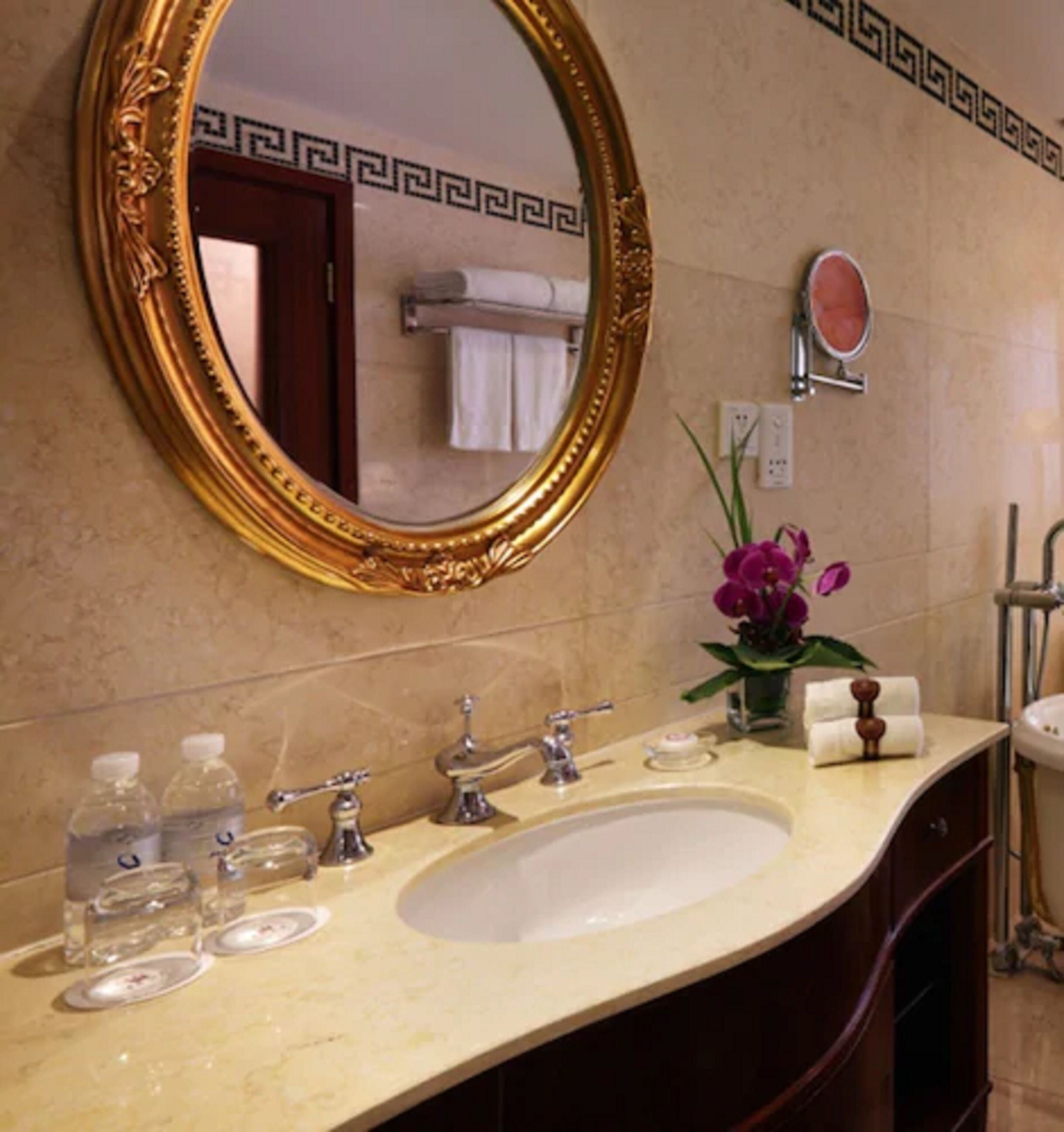 A bathroom with a gold-framed mirror, a sink with two faucets, and a vase with purple flowers.