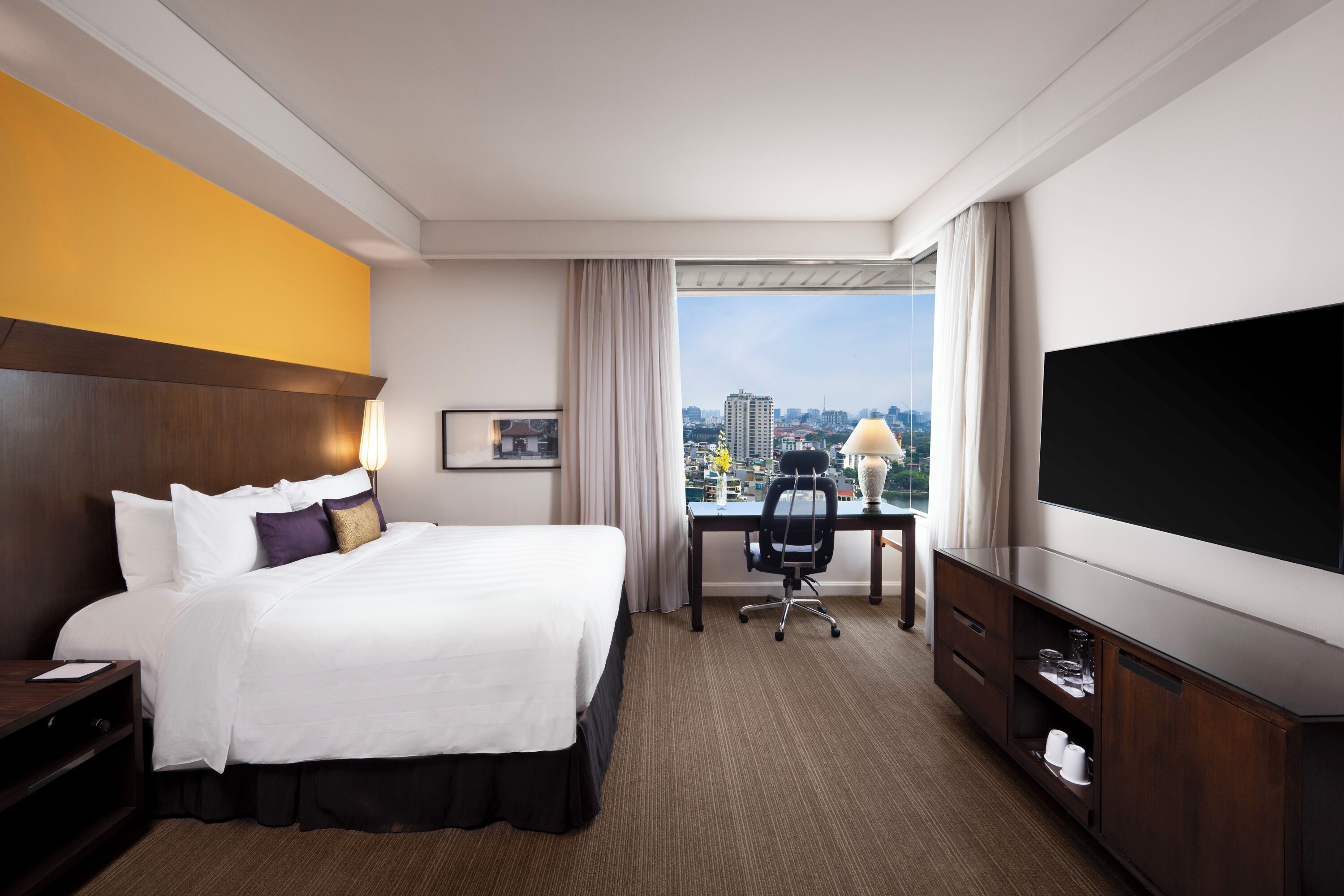 A hotel room with a large bed, a desk, a chair, a TV, and a view of the city.