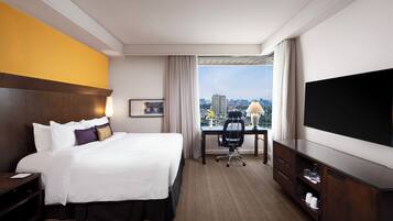 A hotel room with a large bed, a desk, a chair, a TV, and a view of the city.