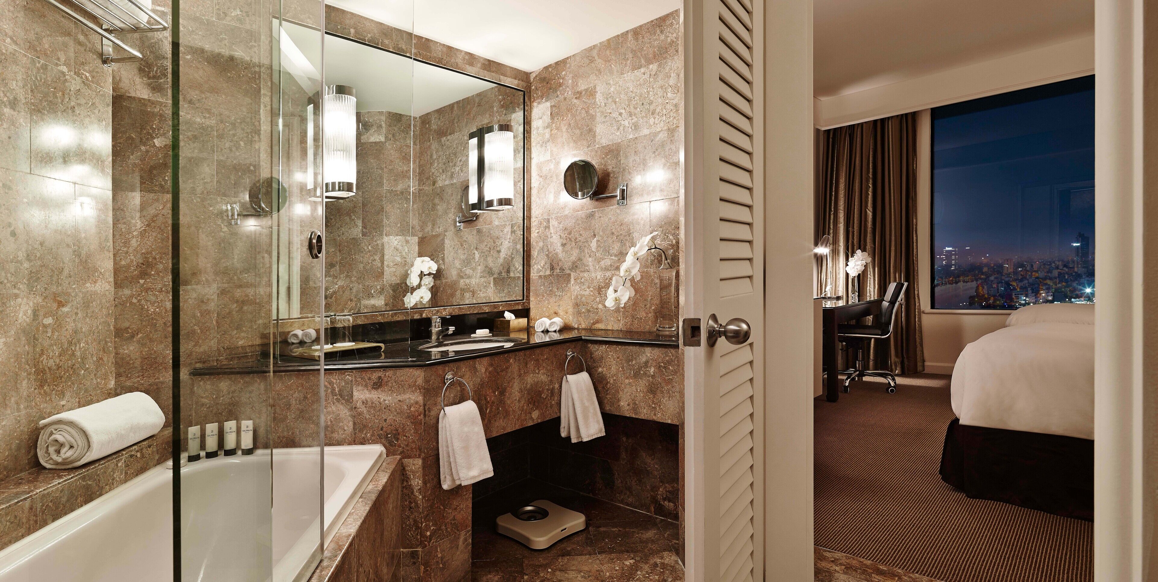 Un moderno baño de hotel con una bañera, ducha con mampara de vidrio, tocador con espejo y un ventanal con vistas a la ciudad.