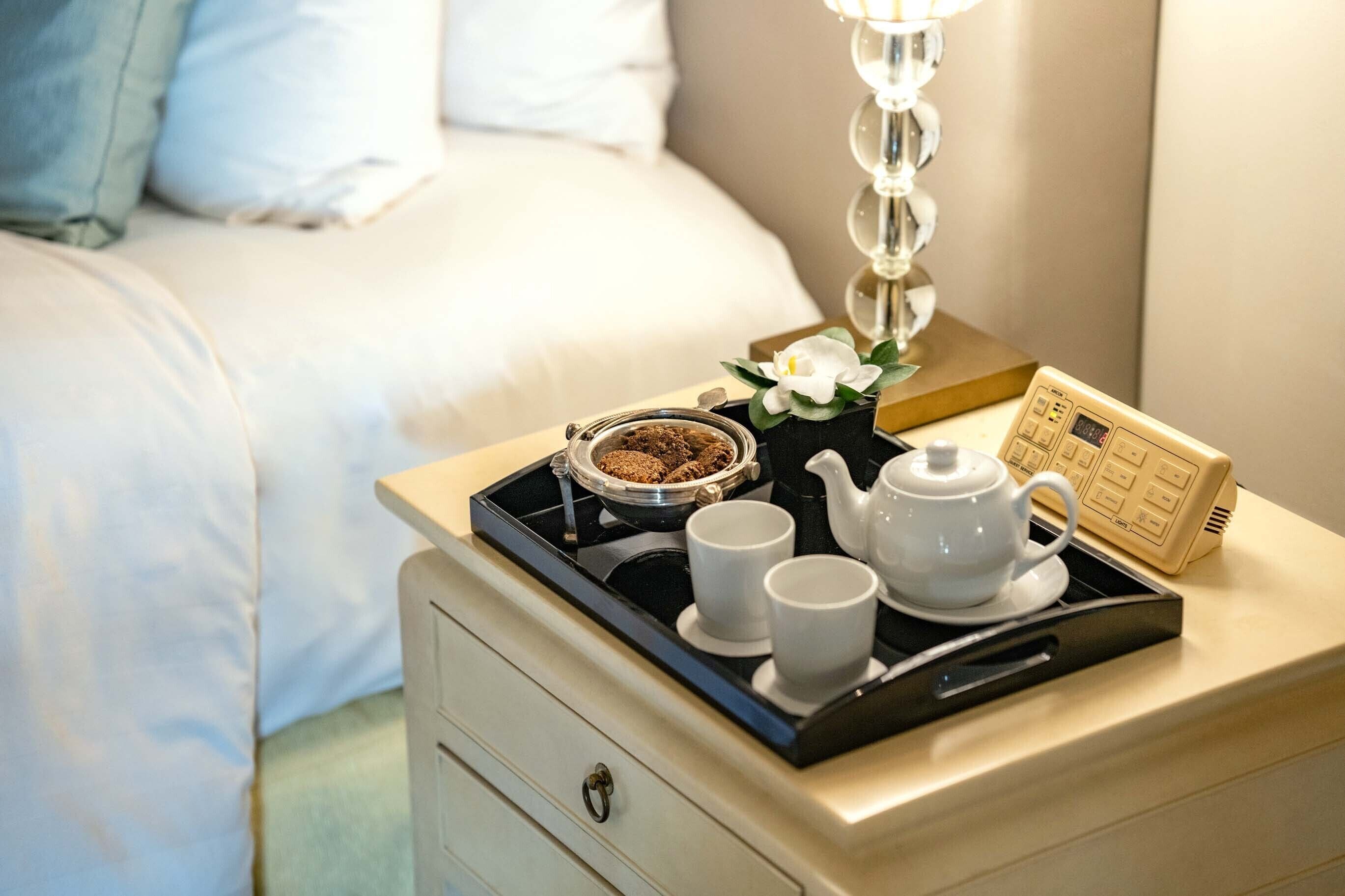 Un lit bien fait, avec du linge de lit blanc, une table de chevet sur laquelle se trouve un plateau avec une théière, des tasses et une petite plante.
