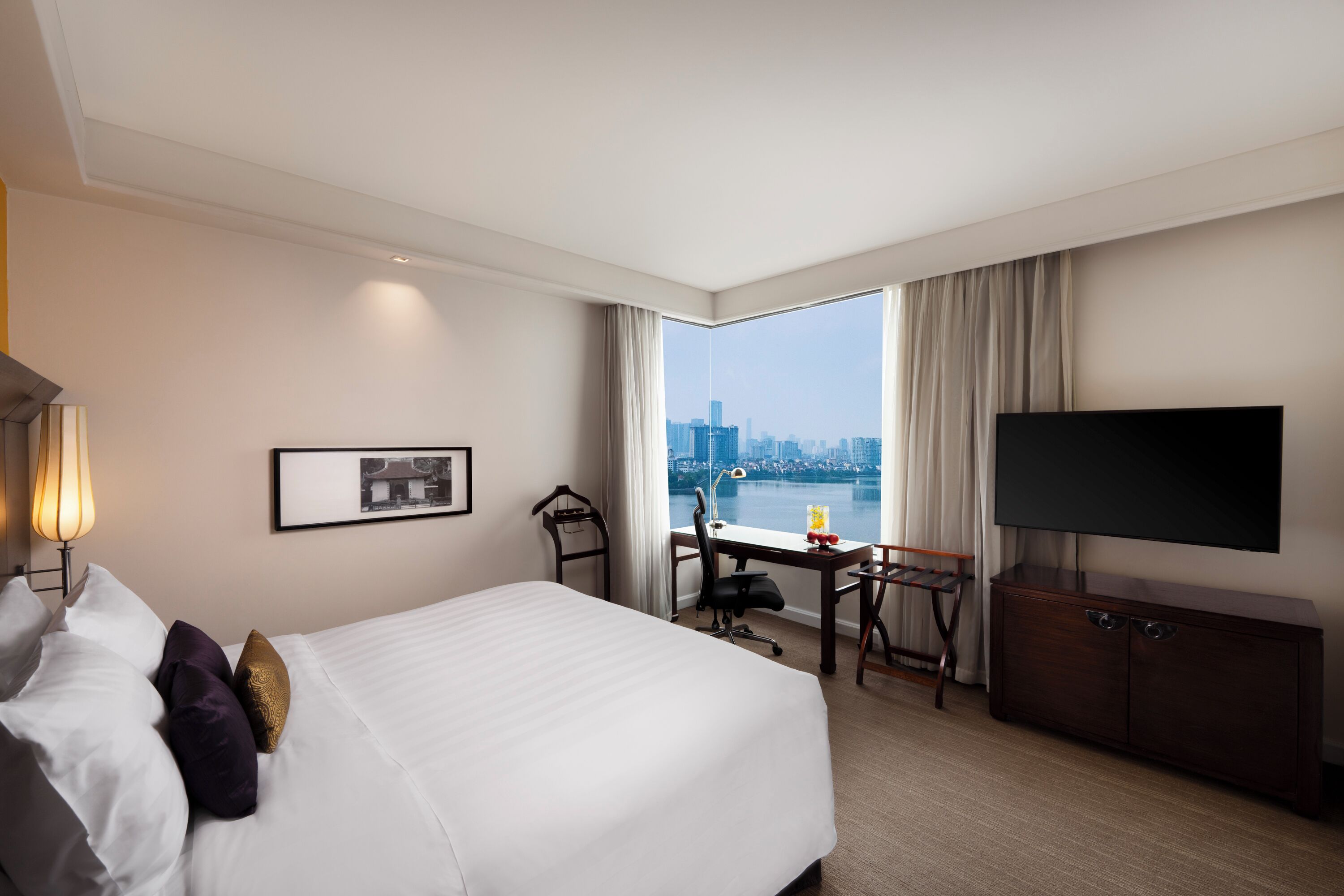 A hotel room with a large bed, a desk, a chair, a TV, and a view of a cityscape.