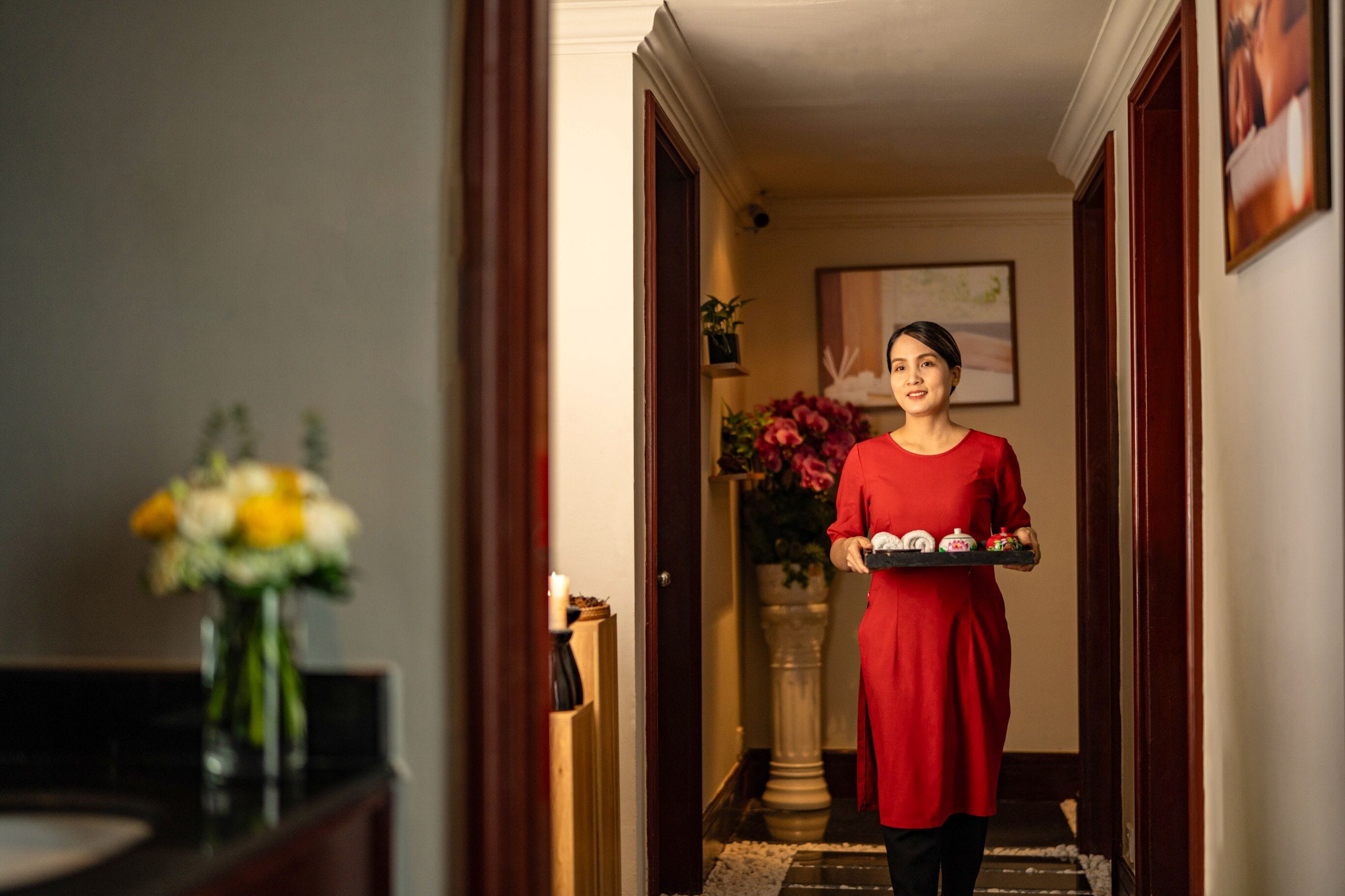 A person in a red dress carrying a tray with items, walking through a hallway with a vase of flowers and a framed picture on the wall.