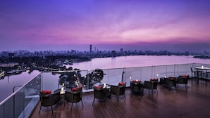 A rooftop terrace with wicker furniture, a view of a city skyline, and a body of water. - Pan Pacific Hanoi (Hanoi)