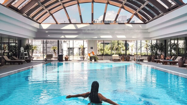 A large indoor swimming pool with a glass ceiling, surrounded by lounge chairs and potted plants. - Pan Pacific Hanoi (Hanoi)