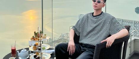 A person seated on a wicker chair by a table with a tea set and pastries, overlooking a body of water with a city skyline in the background.