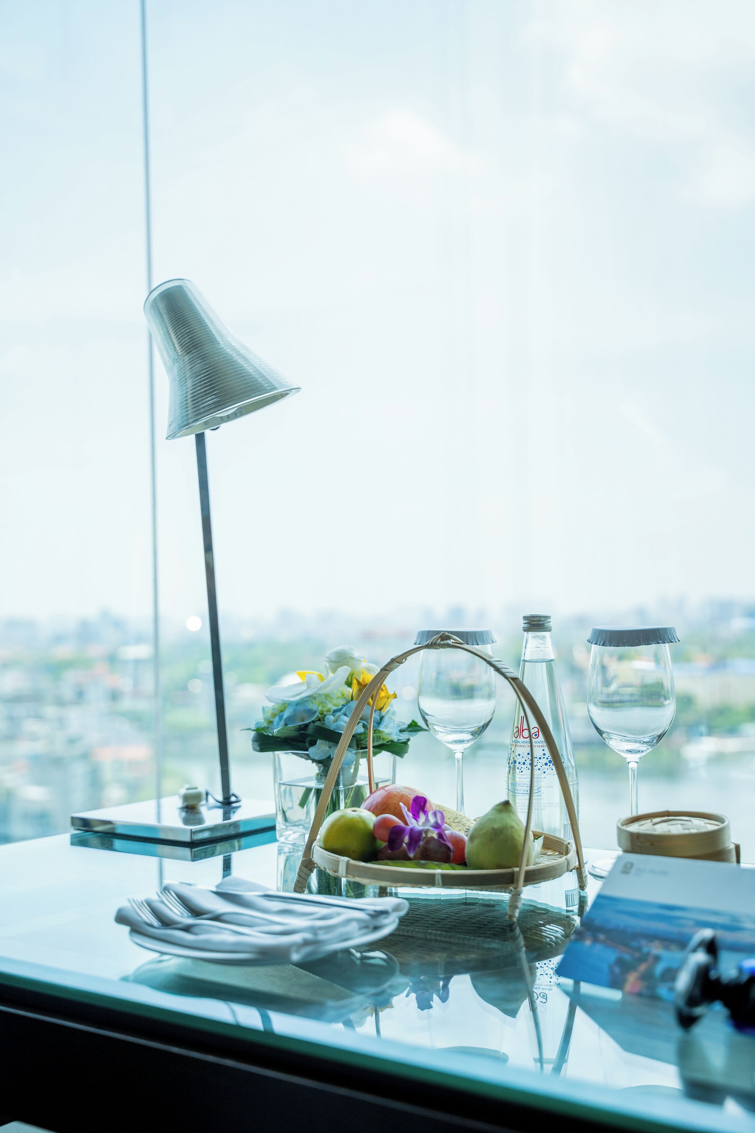 A table with a basket of fruit, two wine glasses, a bottle, and a lamp.