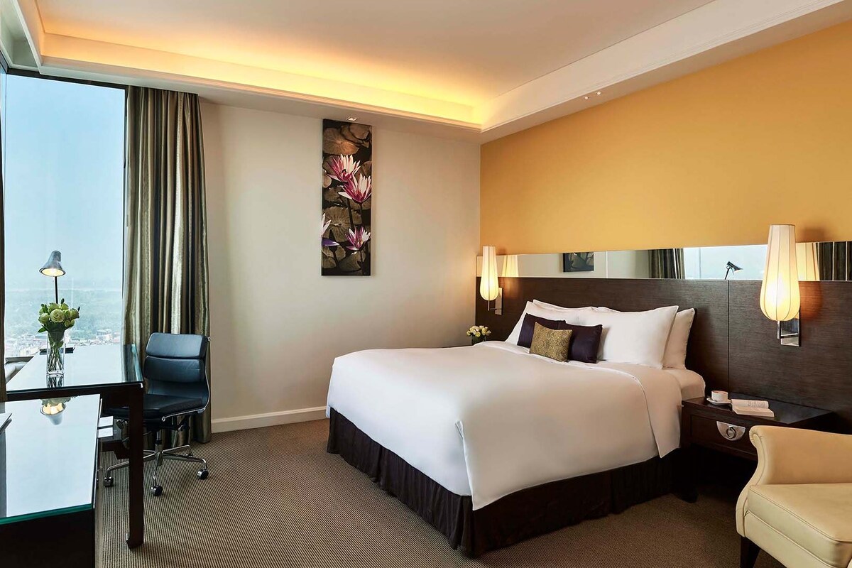 A hotel room with a large bed, a desk with a lamp, a chair, and a view of a cityscape through a large window.