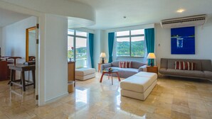 A spacious living room with a sofa, ottoman, and a small coffee table. There is a dining area with a table and chairs, and a view of the outdoors through large windows. - Waterfront Suites Phuket by Centara (Karon)