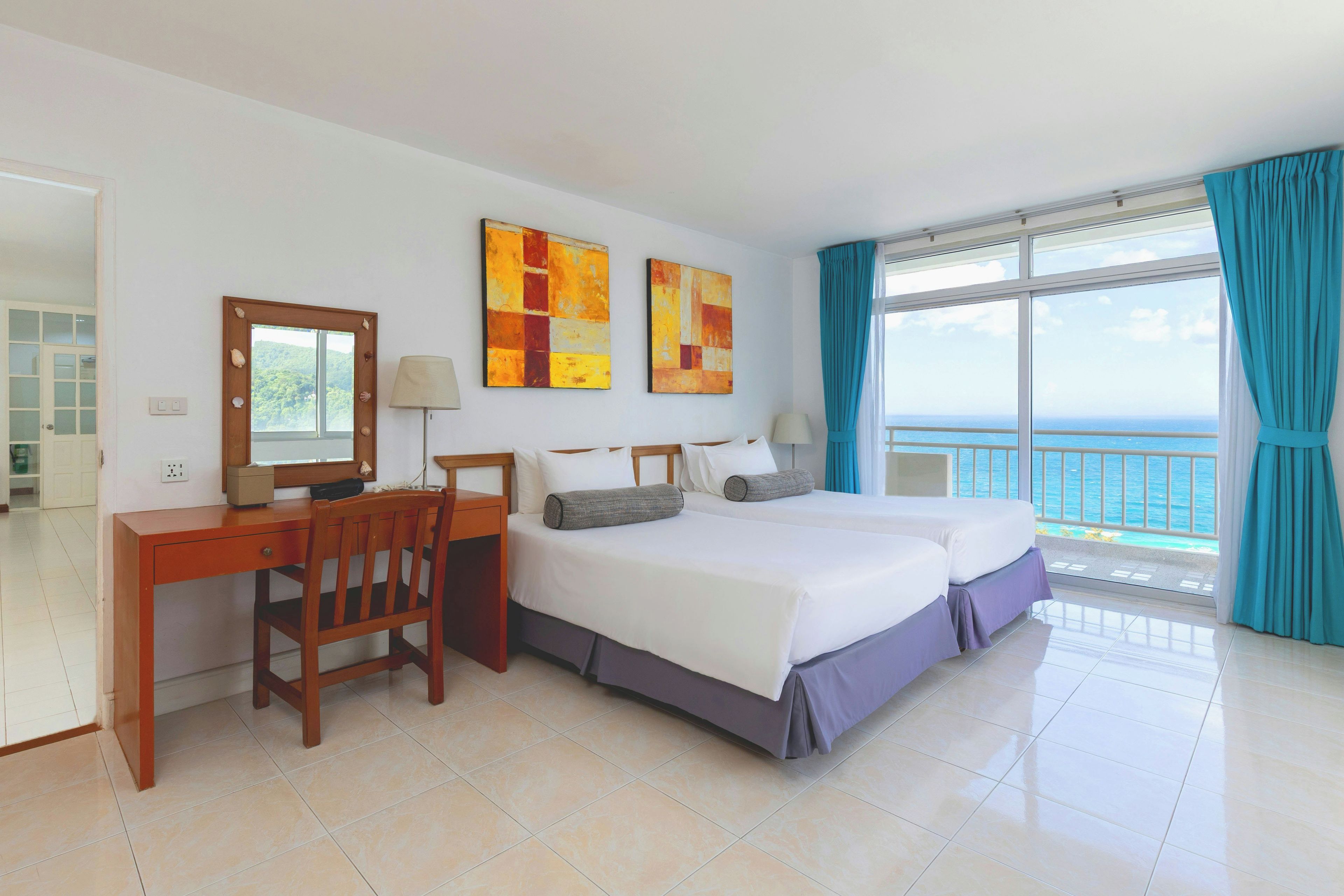 Habitación de hotel con escritorio, dos camas, una silla y un ventanal con vistas al mar.