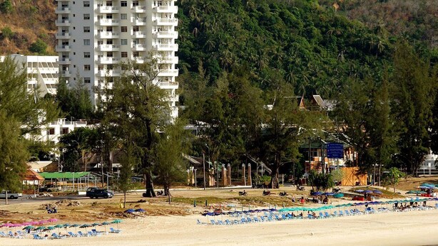 A high-rise hotel with multiple balconies, a beach with umbrellas, and a forested hillside in the background. - Waterfront Suites Phuket by Centara (Karon)
