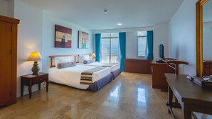 A hotel room with a bed, a desk, a chair, a lamp, a television, and a large window with curtains. - Waterfront Suites Phuket by Centara (Karon)