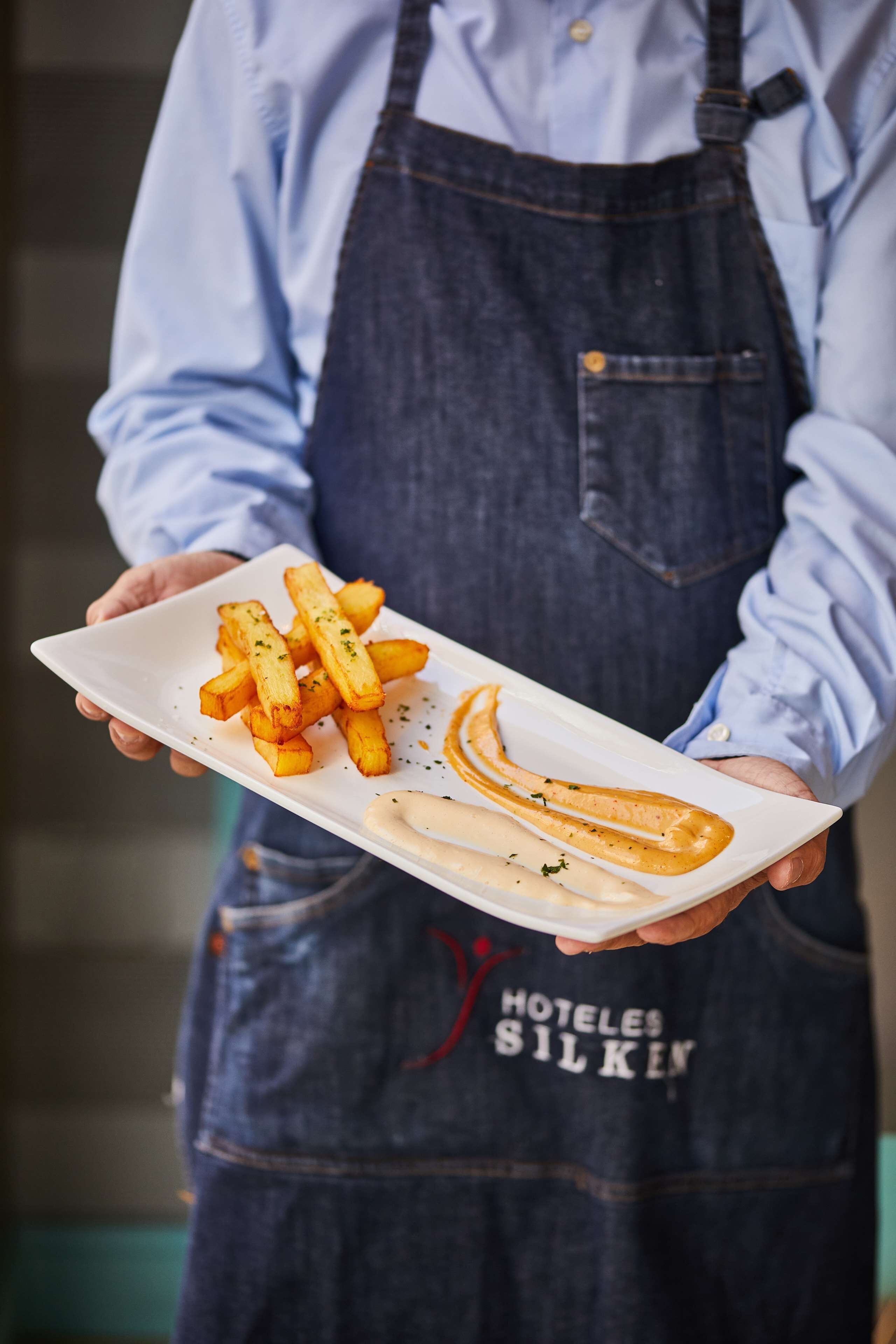 A person holding a white plate with French fries and a sausage.