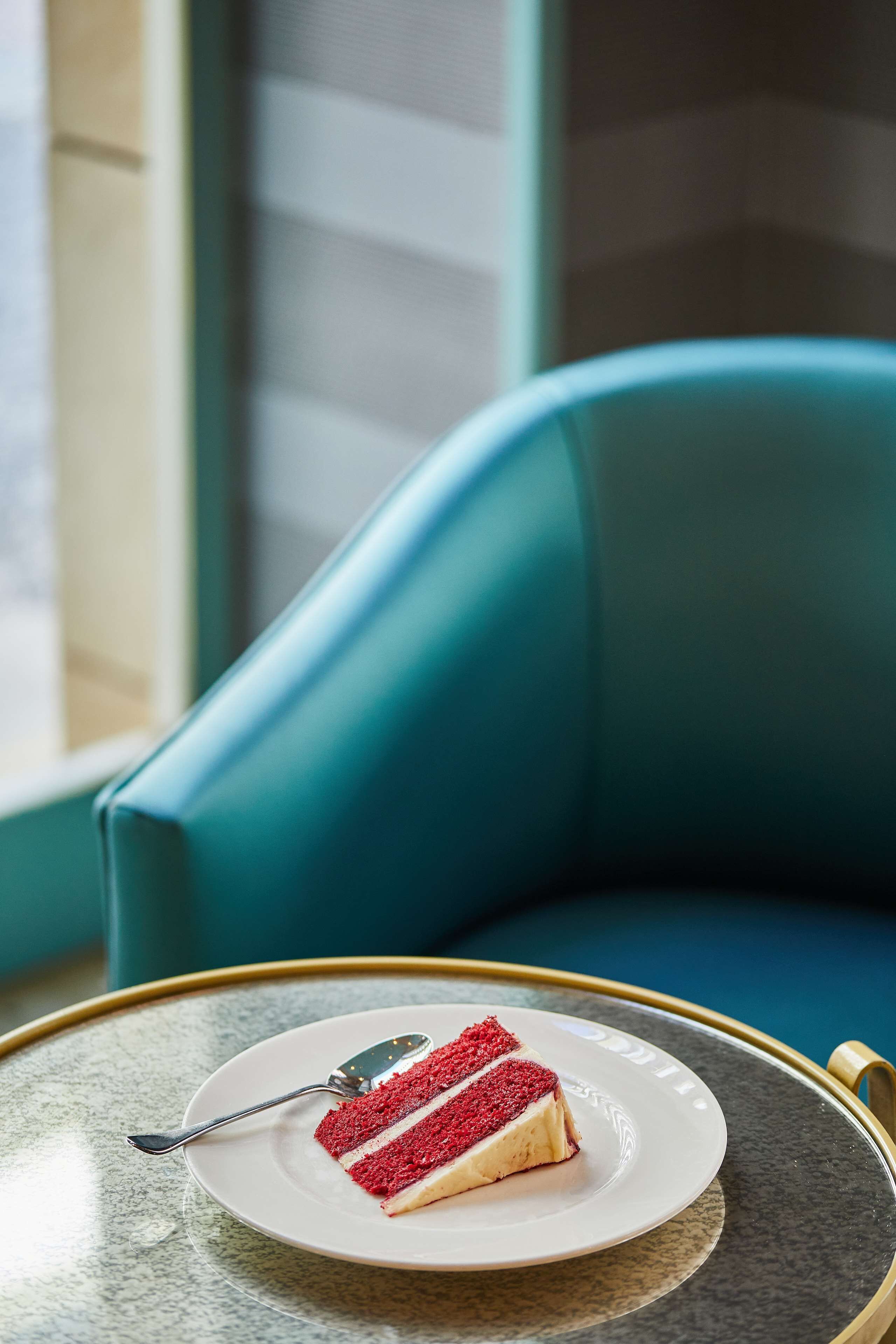 A slice of red velvet cake on a white plate with a spoon, placed on a round table with a gold rim, in front of a teal upholstered chair.