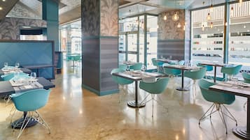 A modern restaurant interior with teal chairs and tables, large windows, and a central column.