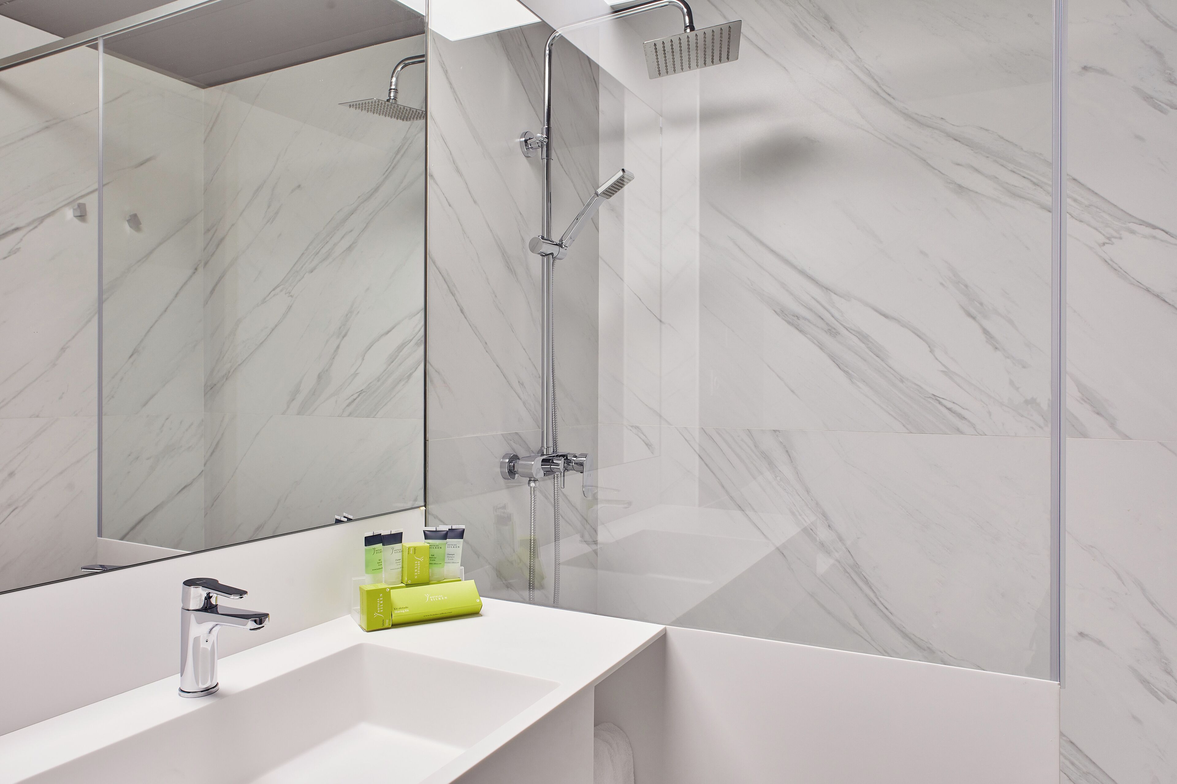 Une salle de bain moderne avec un mur en marbre blanc, un grand miroir, un lavabo blanc et une douche équipée d’un pommeau de douche à main.