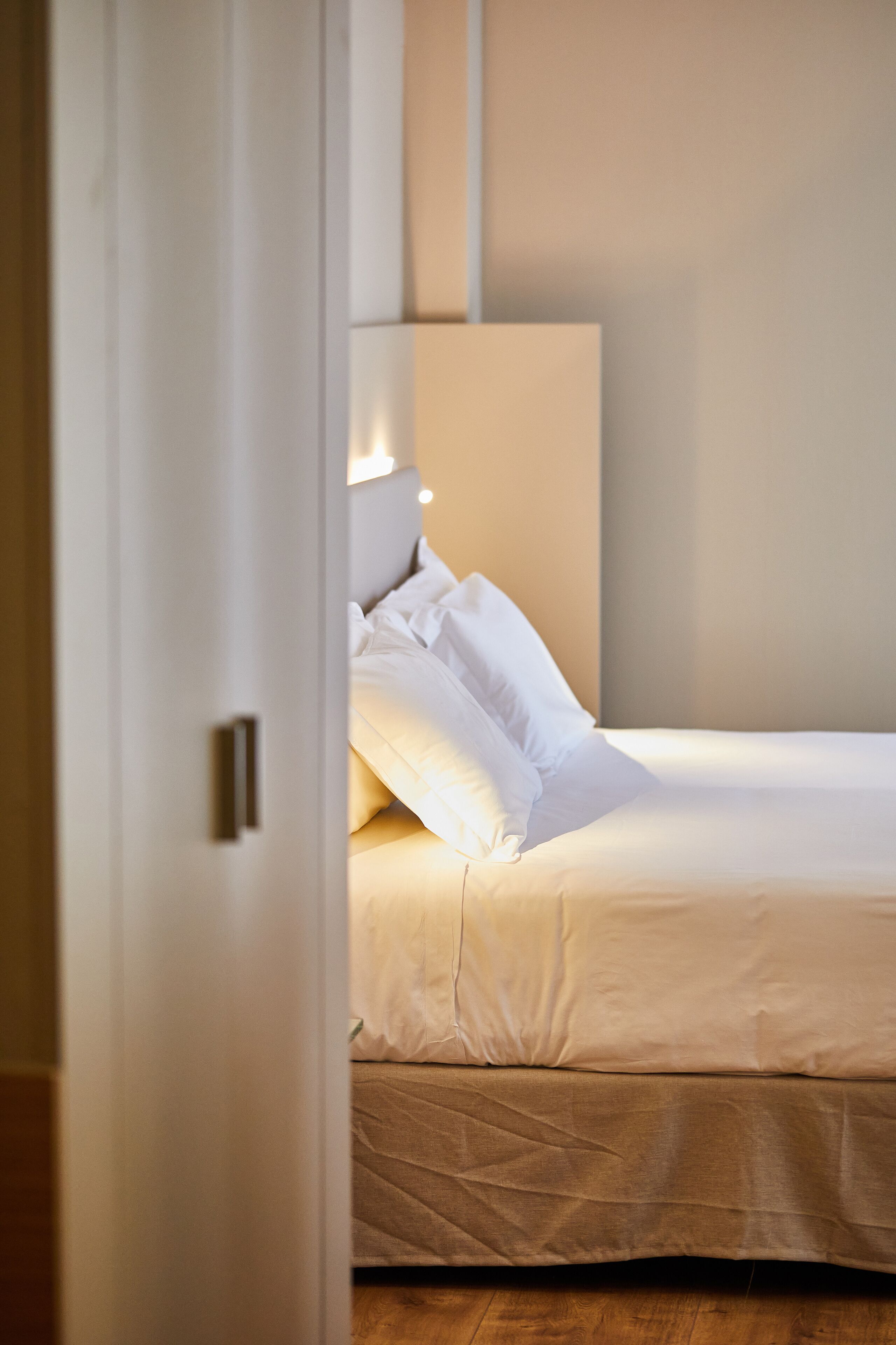 Una cama bien tendida con sábanas blancas y un cabecero, un suelo de madera, y una puerta que da a otra habitación.