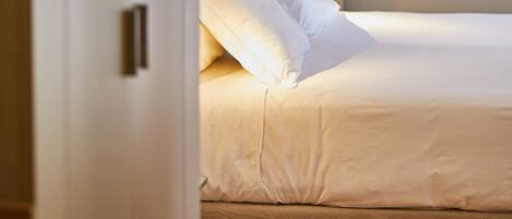 Una cama bien tendida con sábanas blancas y un cabecero, un suelo de madera, y una puerta que da a otra habitación.