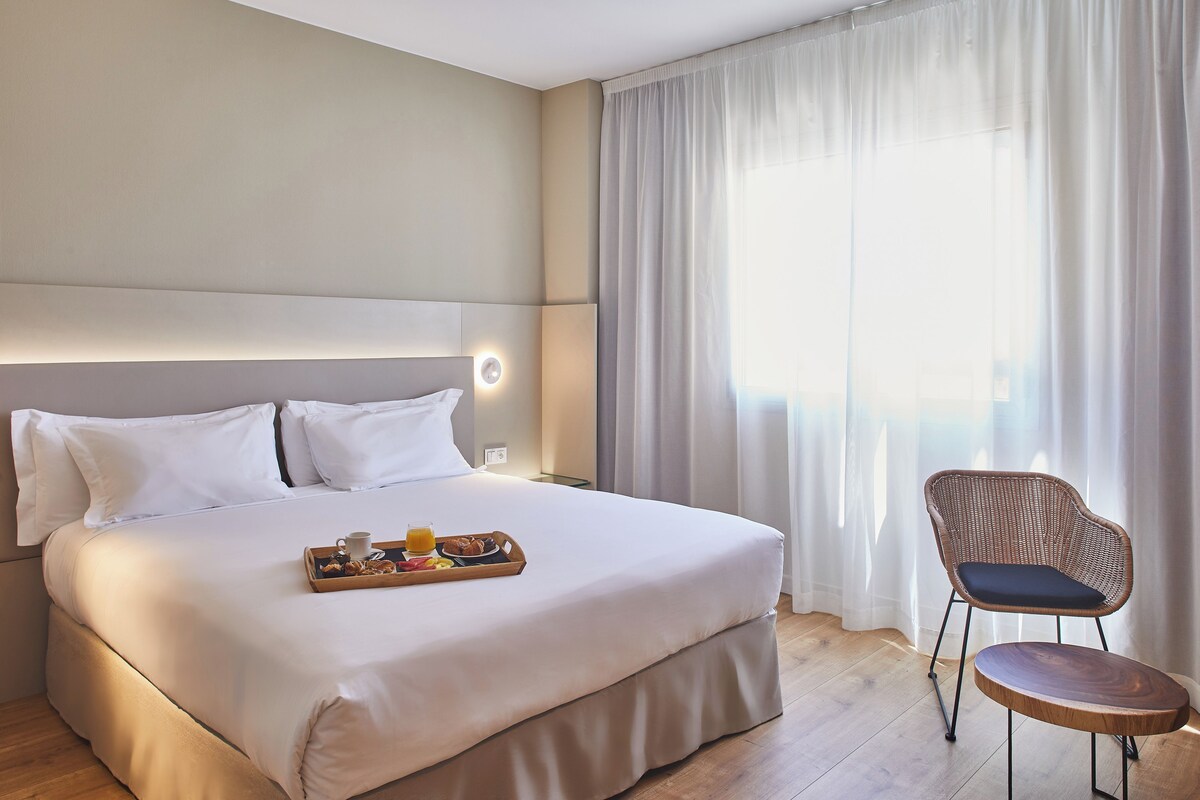 Une chambre d'hôtel avec un grand lit, une table de chevet sur laquelle se trouve un plateau de petit-déjeuner, une chaise et une fenêtre avec des rideaux.