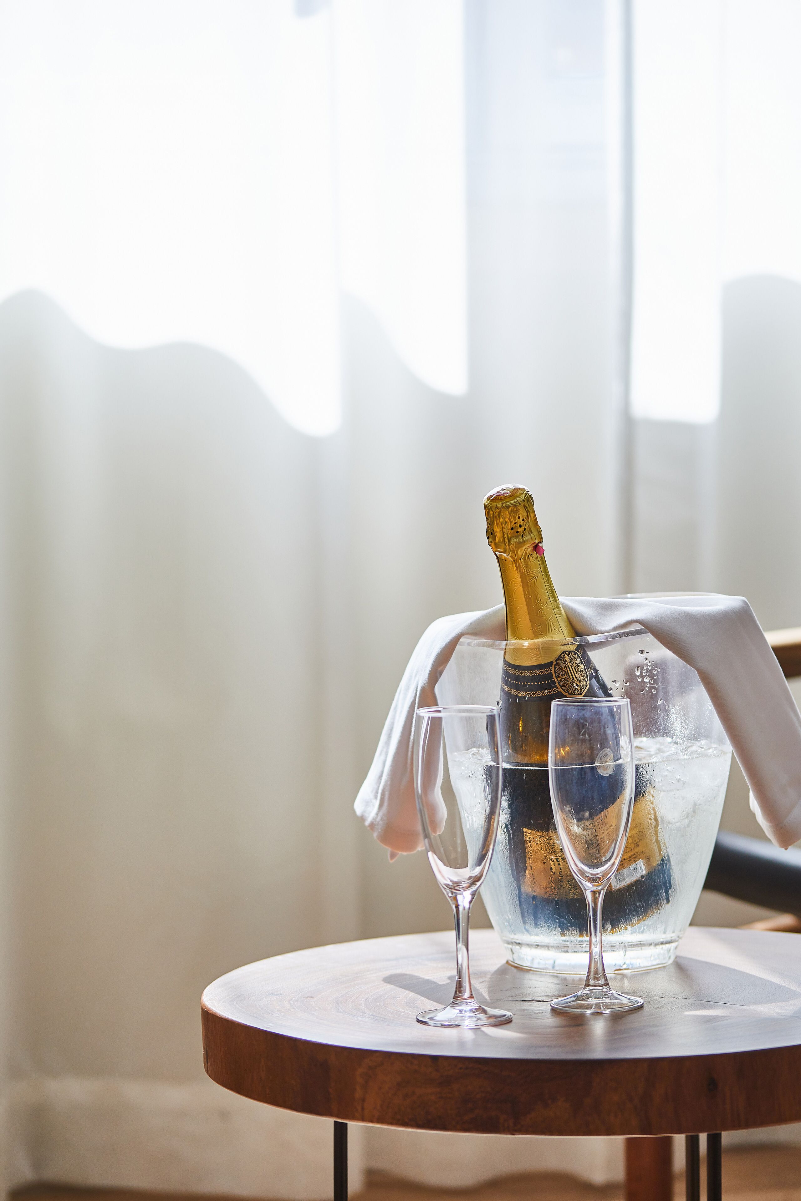 A bottle of champagne in a glass bowl on a wooden table with two glasses beside it.