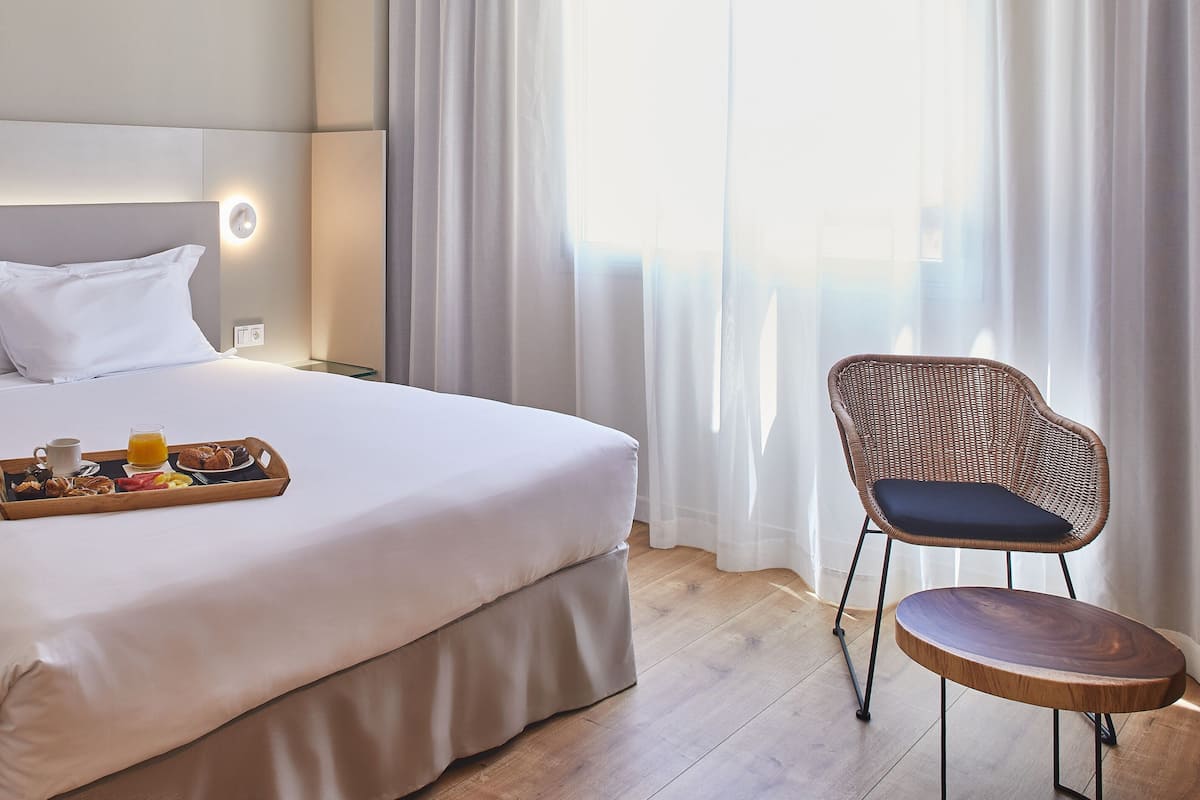 A neatly made bed with a tray of breakfast items, a wicker chair, and a large window with sheer curtains.