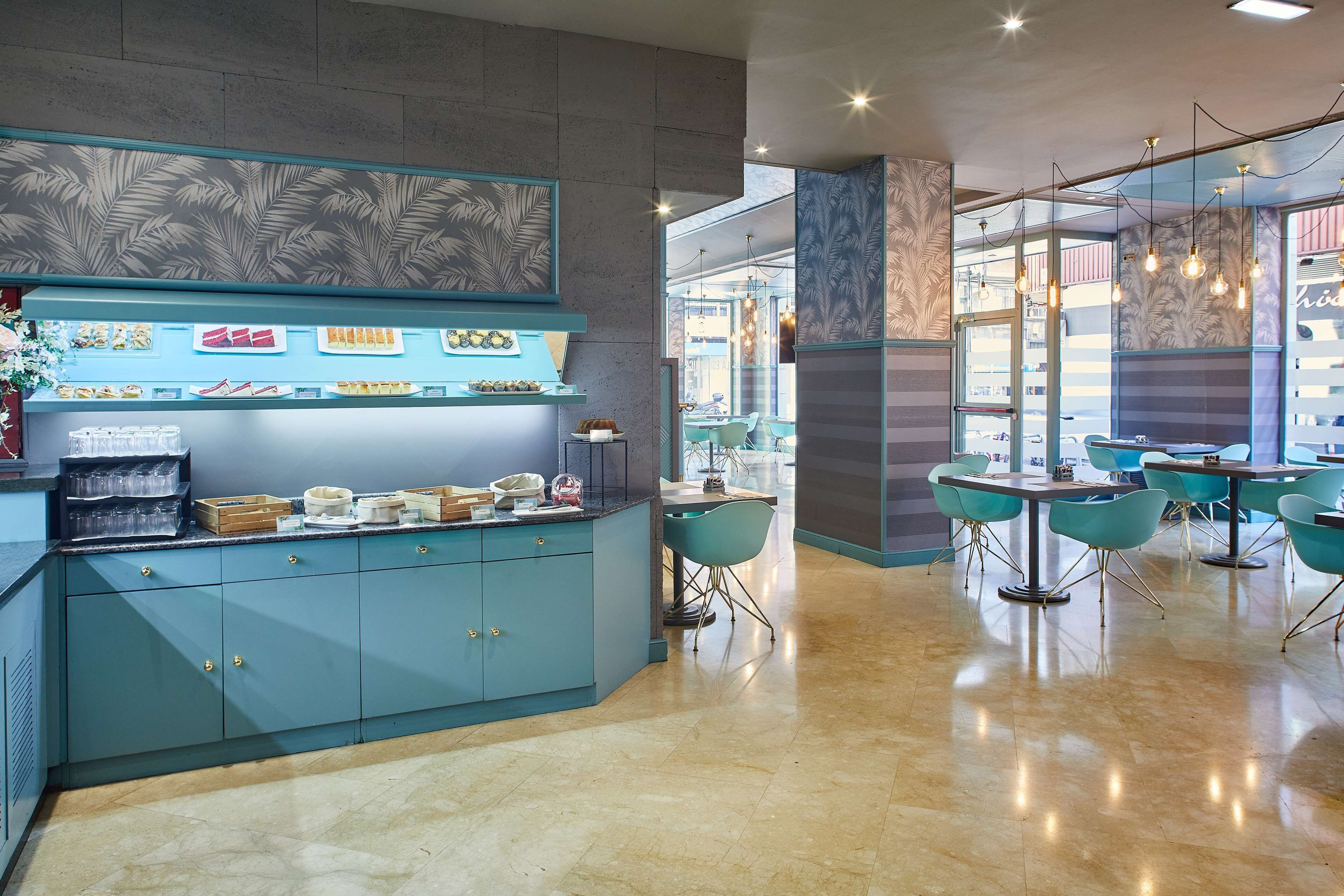 A modern cafe with teal cabinetry, a counter displaying food items, and teal chairs around tables.