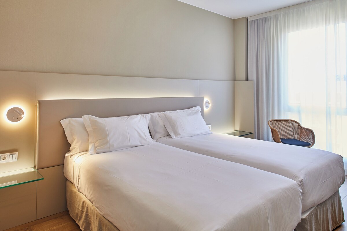 Une chambre d’hôtel avec deux lits, une table de chevet, une chaise et une fenêtre avec des rideaux.
