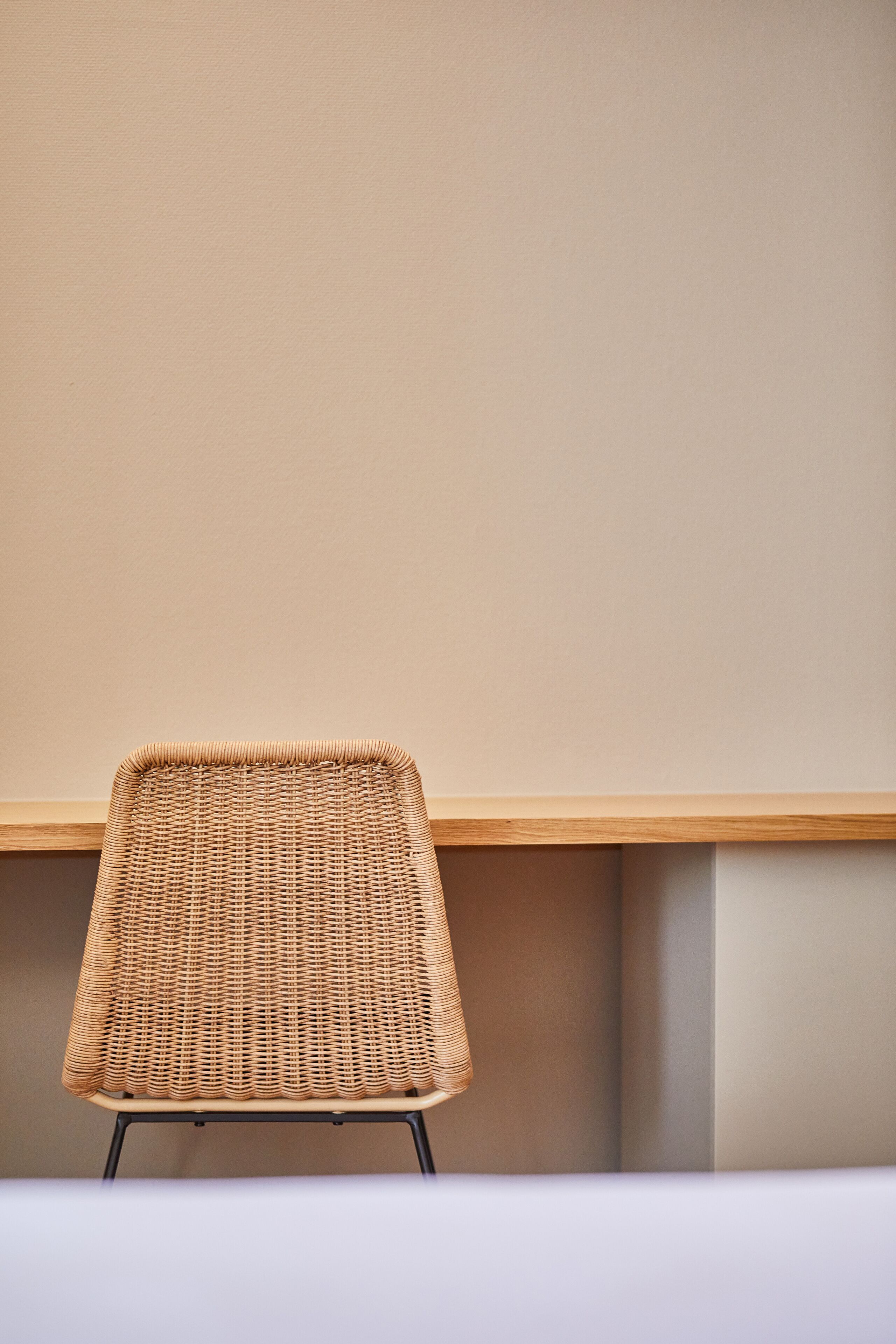 Una silla de mimbre con estructura de madera y patas metálicas, ubicada frente a una pared lisa con un estante de madera en la parte superior.