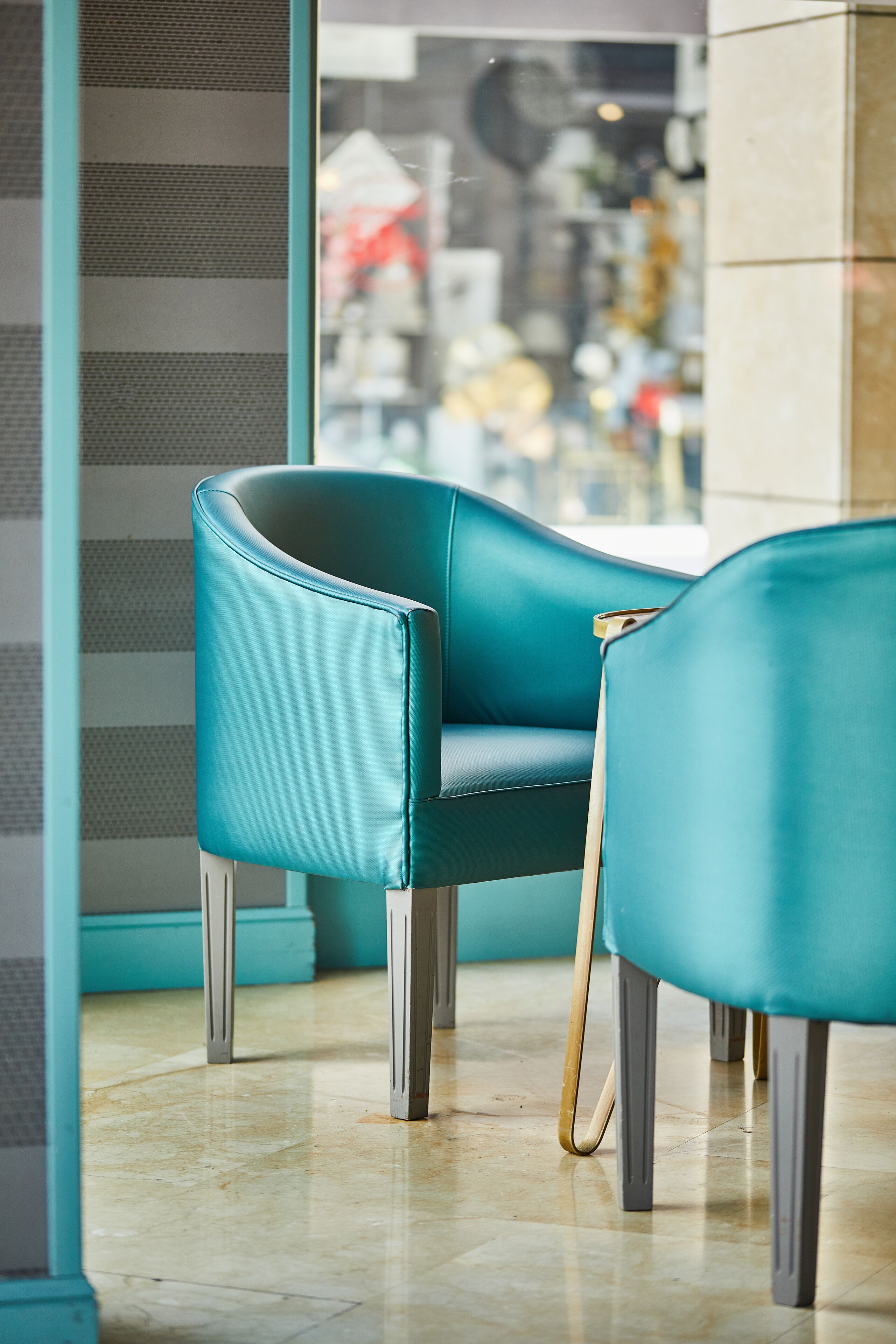 A teal chair with a wooden armrest and metal legs in a room with a tiled floor and a window with a view of a cityscape.