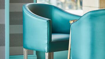 A teal chair with a wooden armrest and metal legs in a room with a tiled floor and a window with a view of a cityscape.