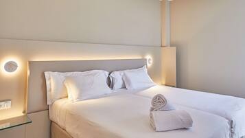 Una cama bien hecha con ropa de cama y almohadas blancas, una mesita de noche de cristal con toma de corriente e iluminación empotrada en la pared.