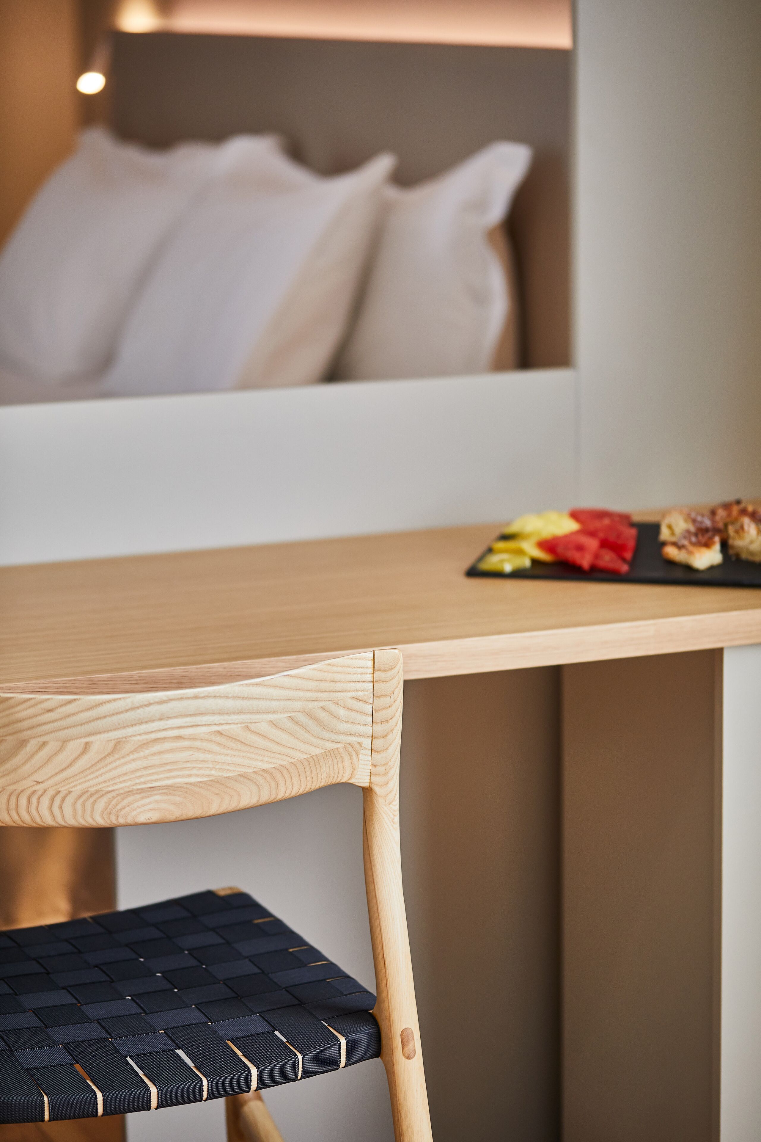 A wooden table with a black woven chair, a white pillow, and a plate of fruit and pastries.