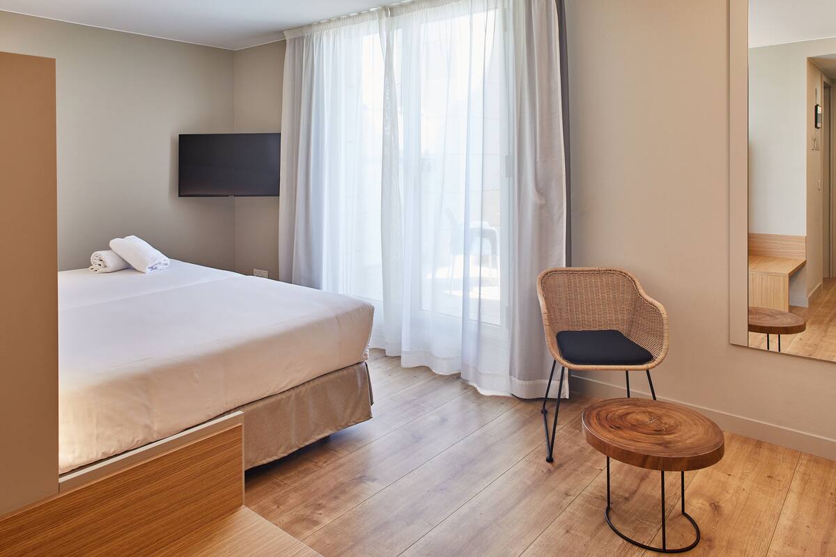 A hotel room with a bed, a chair, a small wooden table, a TV mounted on the wall, and a large window with sheer curtains.