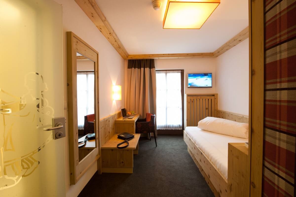Une chambre d’hôtel de taille modeste, avec un lit en bois, un bureau et une chaise, un téléviseur fixé au mur et une fenêtre avec des rideaux.